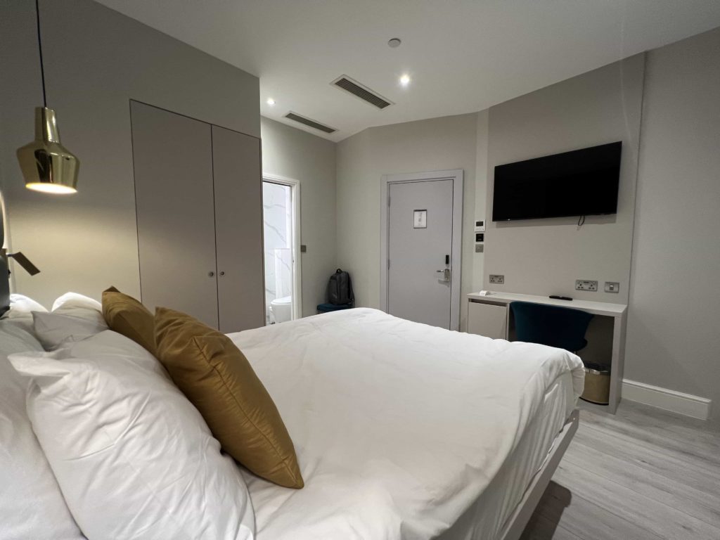 A view from the back corner of a hotel room bed, looking towards a wall-mounted TV