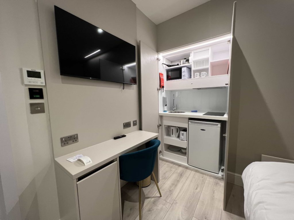 A wall-mounted TV above a narrow desk, with a kitchenette in the background