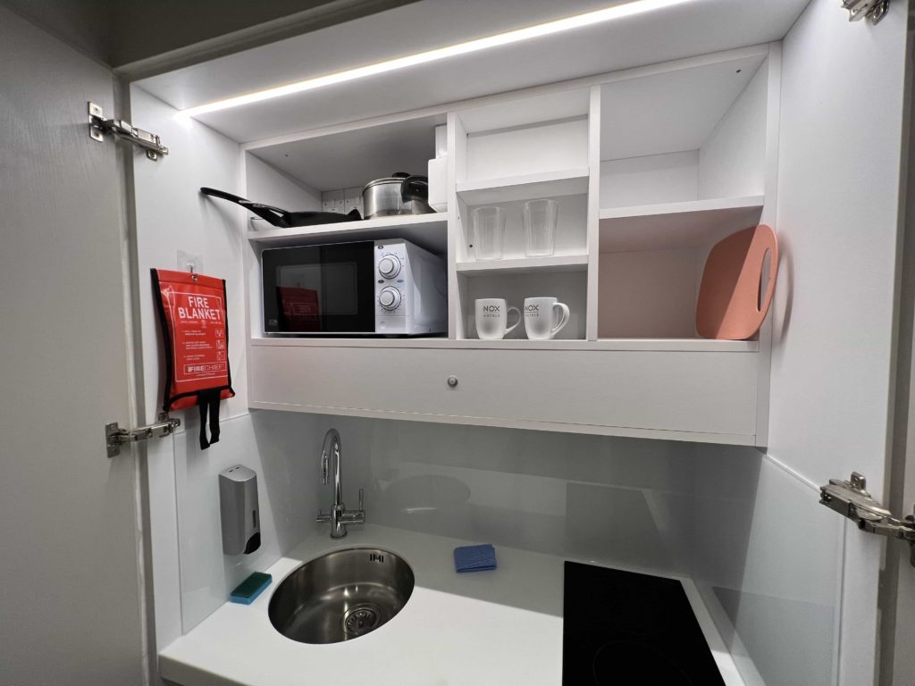 A close-up of a kitchenette countertop which houses a sink and a hob, with shelving above and a microwave