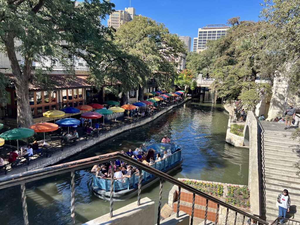 A stretch of the San Antonio riverwalk, with tables shaded under umbrellas and a boat on the river