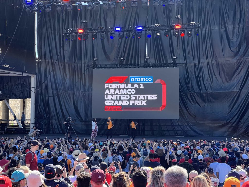 A large crowd in front of an amphitheatre stage, upon which McLaren drivers Lando Norris and Daniel Ricciardo are being interviewed