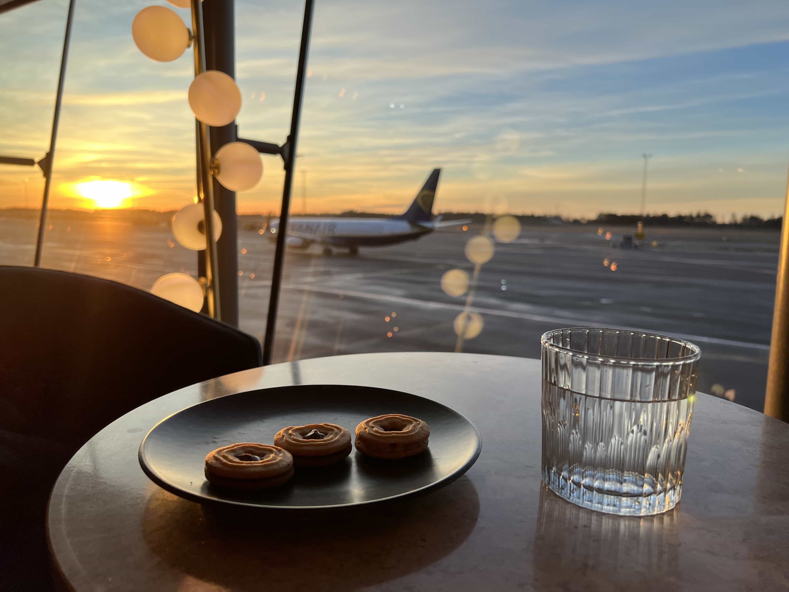 A plate of biscuits, and a glass of tonic water atop a table, with seating that overlooks an airport apron illuminated by the warm glow of the sun