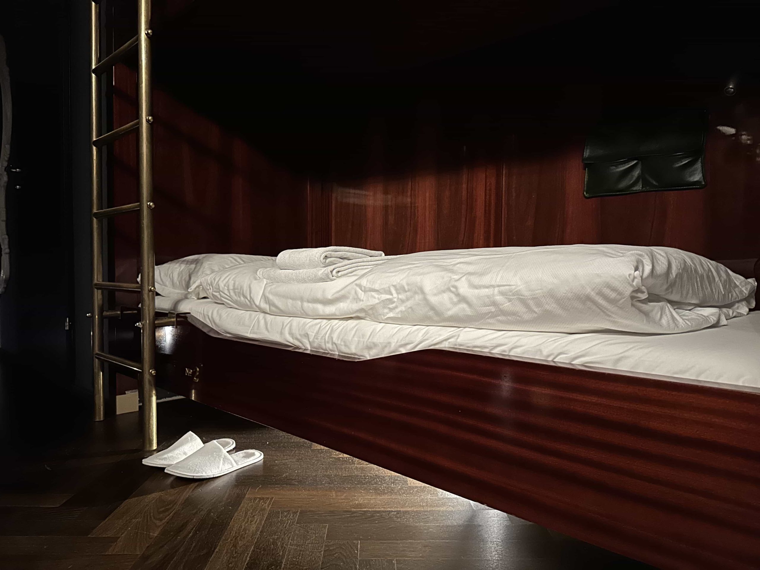 A dorm room bed with neatly folded bedding and towels, with slippers underneath