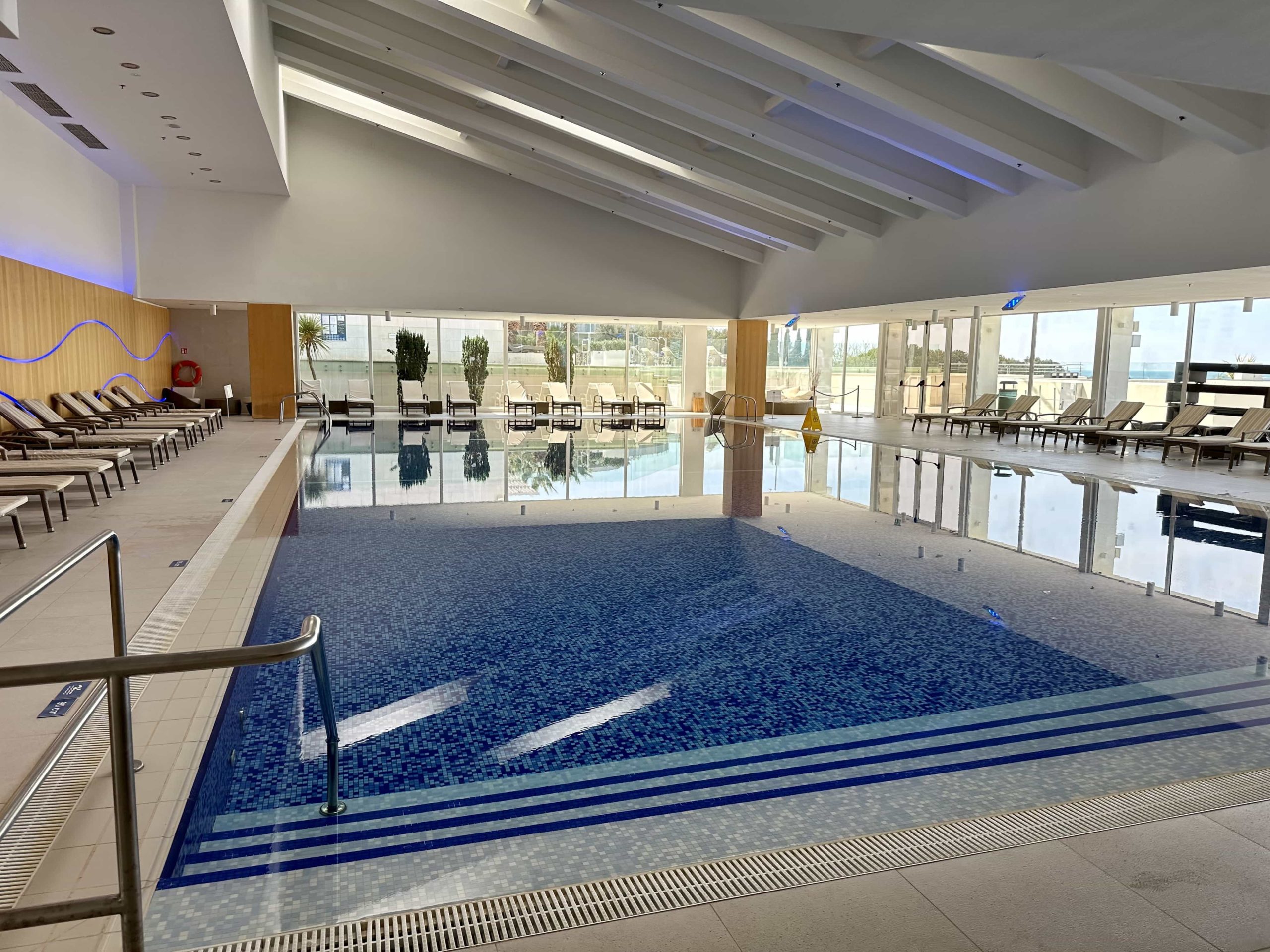 An indoor pool with blue tiles, deck chairs, and wrap-around windows