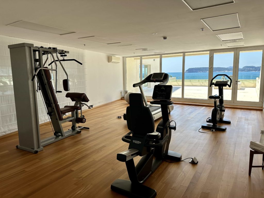 A treadmill, exercise bikes, and a weight machine inside a small fitness room, with windows overlooking the sea